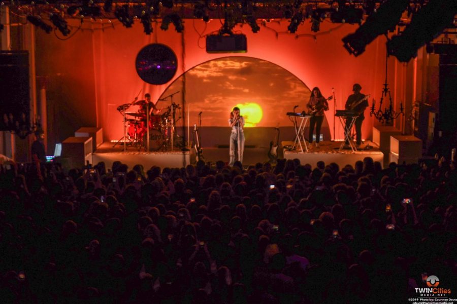 A crowd is gathered in front of Clairo at a concert at the Varsity Theater, the stage has a sunset on the screen behind it